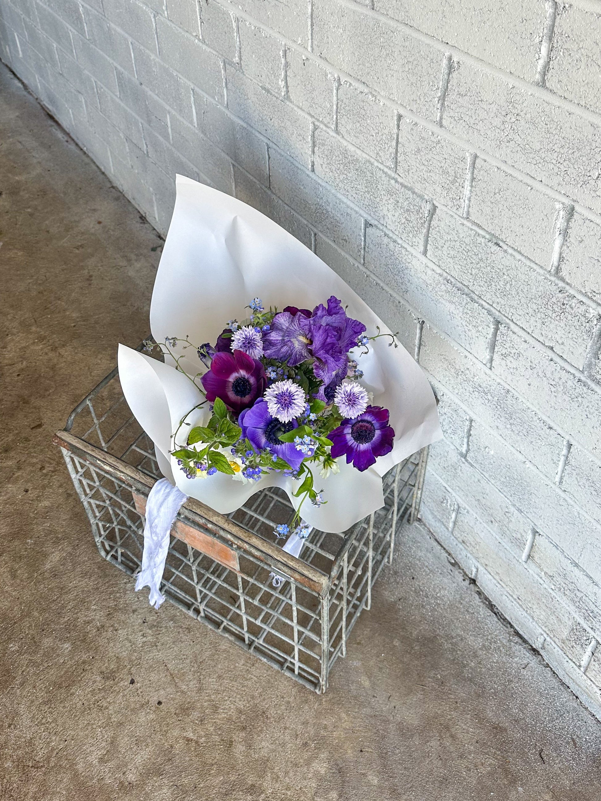Bouquet of purple flowers wrapped in white paper on a wire crate against a gray brick wall.