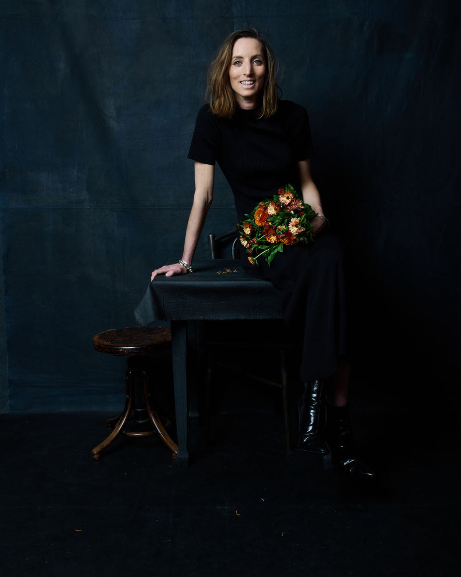 A woman with shoulder-length brown hair sits on a dark table against a dark blue textured backdrop, wearing a black short-sleeve top and black skirt. She holds a bouquet of orange and yellow flowers.
