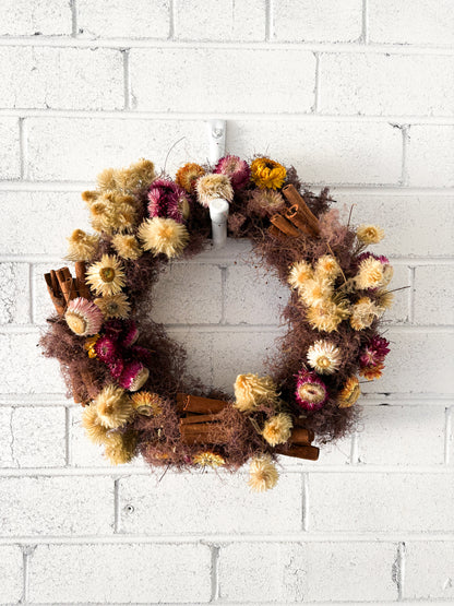 Decorative wreath with dried flowers and cinnamon sticks on a white brick wall.