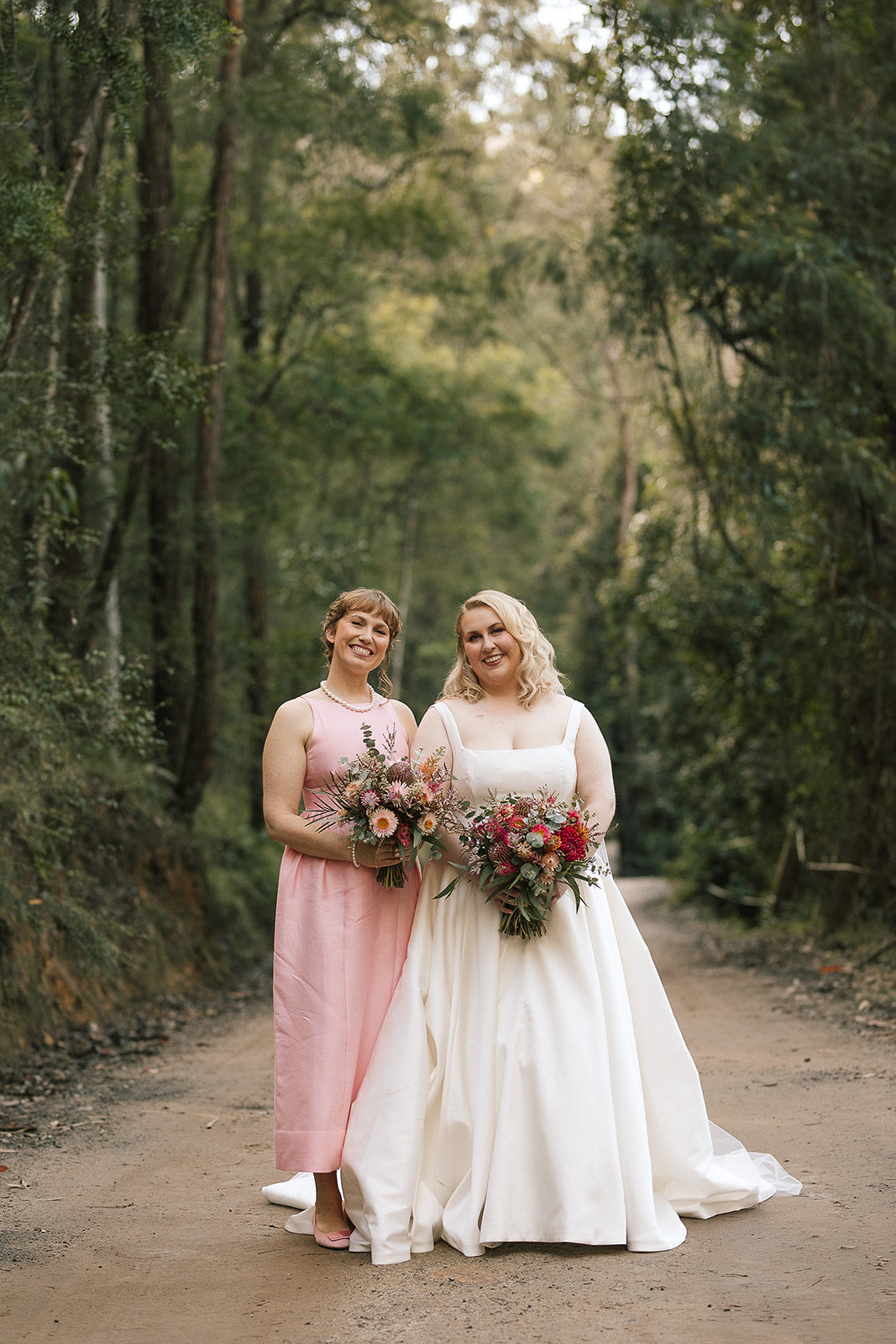 Bride and bridesmaid standing in valley one in a white wedding dress holding flowers, the other in a pink dress.