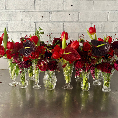 Row of red and burgundy flowers in crystal vases on a metallic surface with a gray brick wall background.