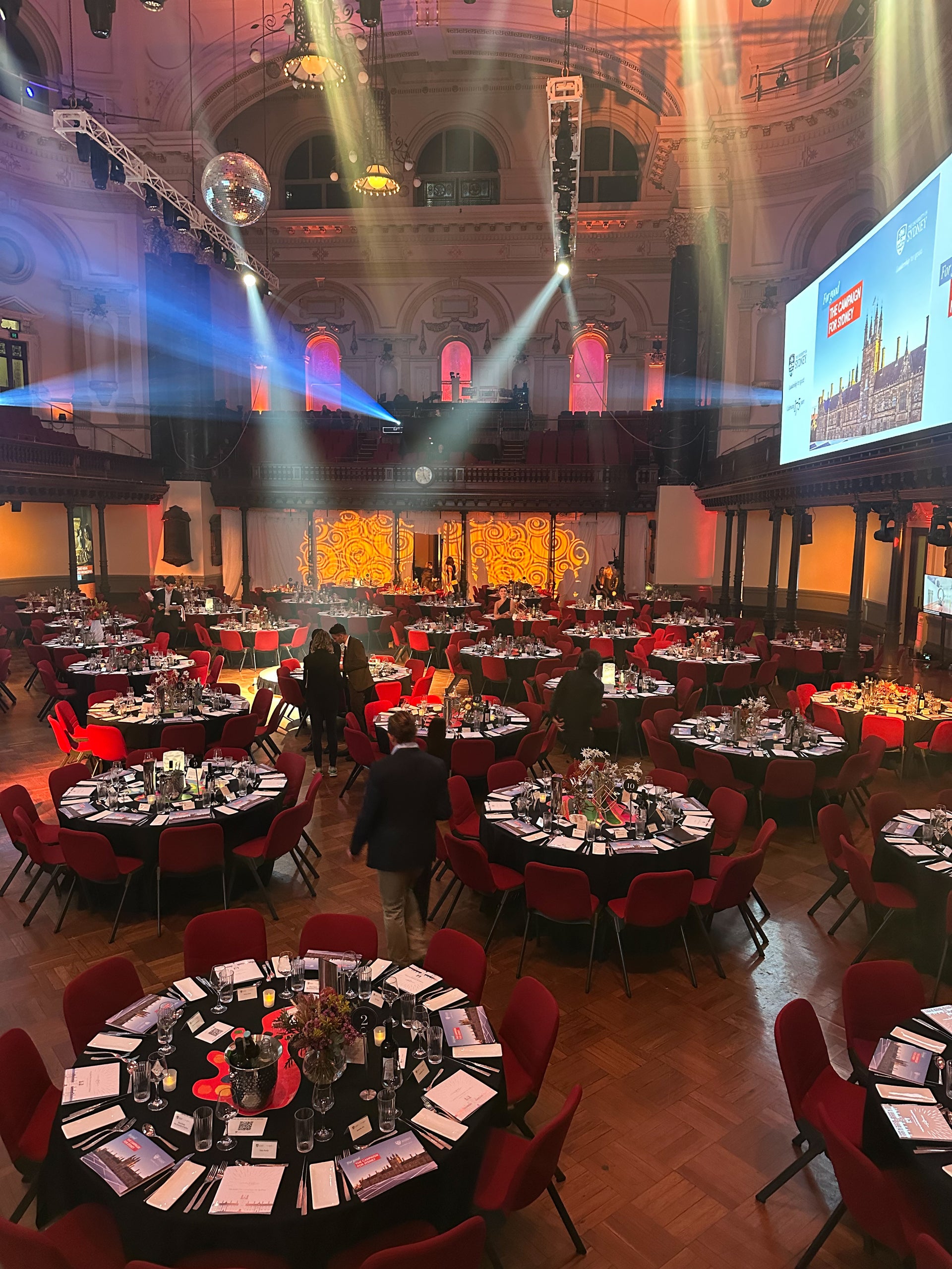 Large banquet hall with round tables, red chairs, and stage setup with lights and screen.